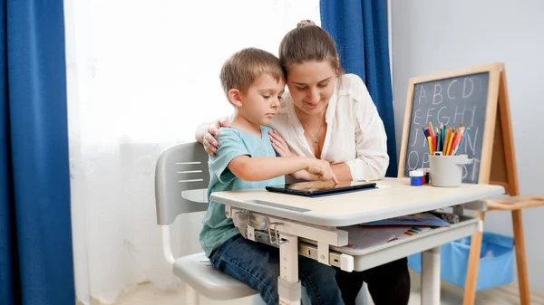 Giovane madre che abbraccia il suo piccolo figlio facendo i compiti e studiando sul tablet a casa. CHild utilizzando gadget per l'istruzione e l'apprendimento — Foto Stock