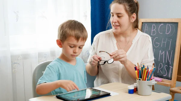 Mutter setzt ihrem smarten Sohn eine Brille auf, spielt am Tablet-Computer und macht Hausaufgaben. Kinderproblem mit Augen und Sehkraft. — Stockfoto