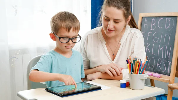 Niño pequeño con anteojos jugando en la tableta. Madre abrazando a su pequeño hijo haciendo la tarea usando gadet. Niños que tienen problemas con los ojos y la vista —  Fotos de Stock