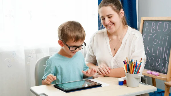Gözlüklü çocuk tablet bilgisayarda oynuyor. Anne küçük oğluna sarılıp ödev yaparken gadet kullanıyor. Çocukların görme ve görme sorunları var. — Stok fotoğraf
