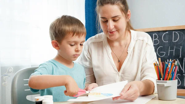 Junge Lehrerin zeigt ihrem kleinen Schüler, wie man mit der Schere umgeht. Junge schneidet Papier mit Schere bei Hausaufgaben. — Stockfoto