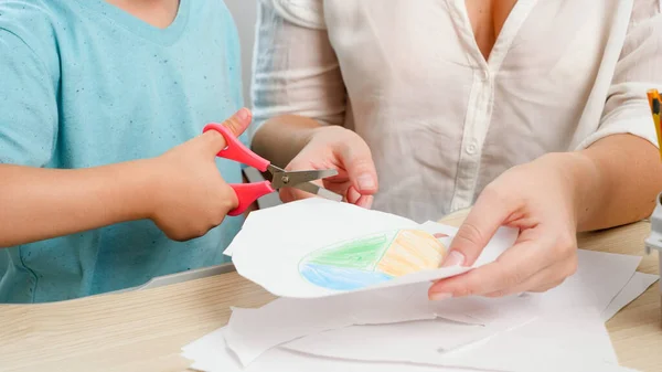 Kleiner Junge schneidet mit der Schere gezeichnetes Bild auf Papier aus. Mutter hilft ihrem kleinen Sohn. Bildung und Lernen zu Hause — Stockfoto