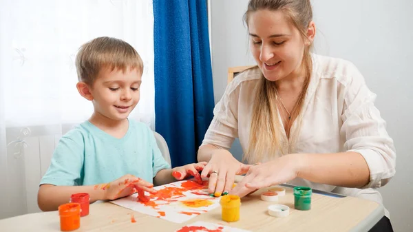 Szczęśliwy uśmiechnięty chłopiec z brudnymi dłońmi w kolorowej farbie patrząc na rysunek matki z palcami i gwasz. rodzina zabawy razem i tworzenia rysunków artystycznych. — Zdjęcie stockowe