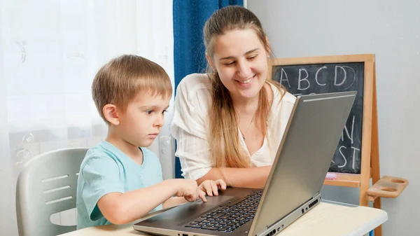 Lachende moeder die haar zoontje les geeft met laptop tijdens het onderwijs en thuis. Begrip afstandsonderwijs en afstandsonderwijs thuis — Stockfoto