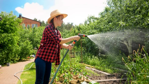 Young smiling girl watering garden from garden hose on hot summer day. Woman watering growing plants and working at backyard — Stock Photo, Image