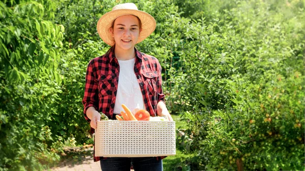 Portret van een vrolijke glimlachende vrouwelijke boer die in de tuin loopt en de box vasthoudt met rijpe biologische groenten. Concept van kleine bedrijven en het telen van biologische groenten in de tuin — Stockfoto