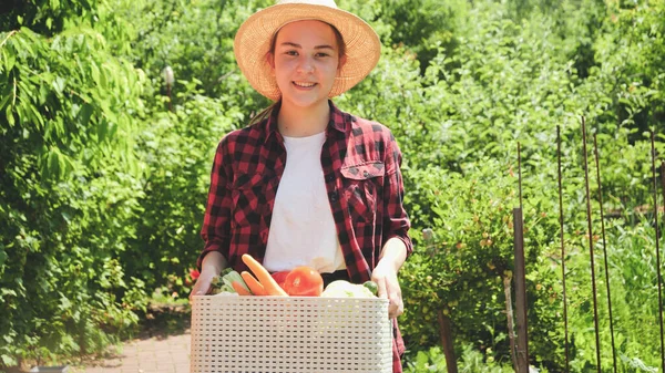 Mujer sonriente en caja de sujeción sombrero con verduras orgánicas maduras y caminando en el jardín. Concepto de pequeña empresa y cultivo de verduras orgánicas en el jardín del patio trasero — Foto de Stock