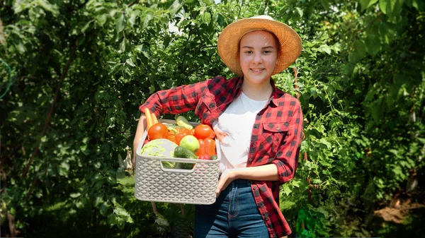 Chica sonriente en sombrero caminando en arden con la cosecha en caja grande. Verduras orgánicas frescas y maduras en caja. Concepto de pequeña empresa y cultivo de verduras orgánicas en el jardín del patio trasero — Foto de Stock