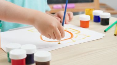 Küçük çocuk masanın arkasında oturuyor ve boya ve fırçalarla renkli resimler çiziyor. Tecrit ve tecrit süresince evde çocuk eğitimi. Sanat kavramı ve çocukların yaratıcılığı
