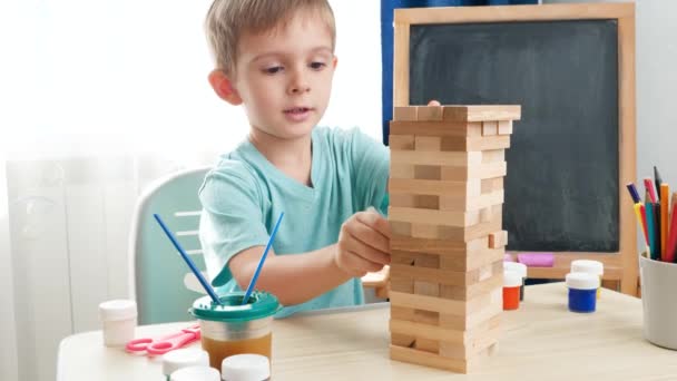 Niño inteligente sacando bloques de madera de la torre alta hecha de ladrillos. Concepto de niños inteligentes y educación en el hogar durante el encierro y el autoaislamiento. — Vídeos de Stock