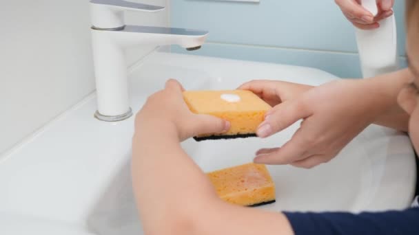 Κοντινό πλάνο του μικρού αγοριού που βοηθάει τη μητέρα να πλύνει το νεροχύτη με σφουγγάρι και απορρυπαντικό στο μπάνιο. Η έννοια της οικογενειακής ομαδικής εργασίας και της οικιακής εργασίας — Αρχείο Βίντεο