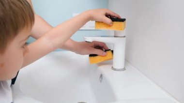Sünger ve deterjanla lavabo ve su musluğunu temizleyen küçük çocuğun yakın çekimi. Hijyen ve ev işi kavramı. Daiy rutininde ebeveynlere yardım eden çocuklar