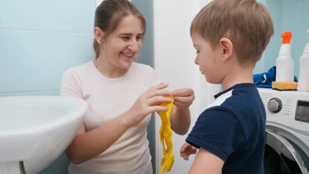 Leende ung mor hjälpa lille son att sätta på gula gummihandskar innan du gör hushållsarbete i badrummet. Barn som hjälper föräldrar med sysslor och dagliga rutiner. — Stockvideo