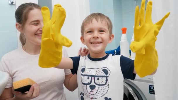 Смішний хлопчик, який сміється в жовтих гумових рукавичках, сміється і робить обличчя, перш ніж робити домашню роботу і прибирати ванну кімнату зі своєю матір'ю. Сімейні домашні завдання та розваги — стокове відео