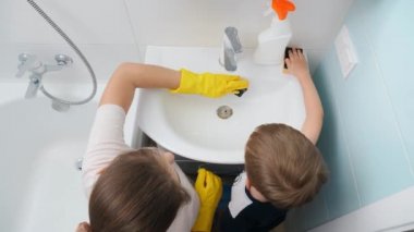 Genç bir anne, küçük oğluna banyoda lavaboyu yıkamayı ve temizlemeyi öğretiyor. CHild, ebeveynlere ev işlerinde yardım ediyor.