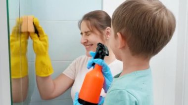 Küçük çocuk spreyden kimyasal deterjan sürerken annesi banyo aynasını ovup temizliyor. Aile ev temizliği ve ev işi yapıyor.