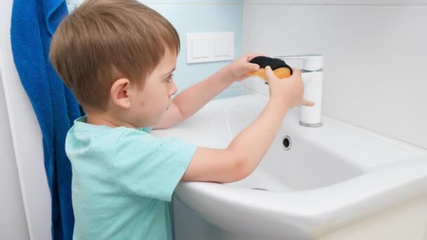 Pequeño niño lavando y limpiando el lavabo en el baño con esponja y detergente o limpiador. — Vídeo de stock