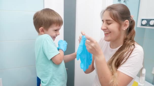 Усміхнена мати розважається зі своїм маленьким сином, одягаючи захисні гумові рукавички перед прибиранням та миттям у ванній кімнаті — стокове відео