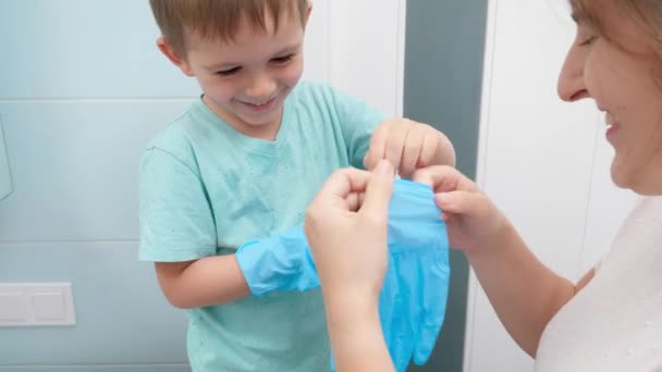 妈妈笑着帮助她的小儿子穿上防护的橡胶手套，然后才打扫房间。家庭做家务活，玩得开心 — 图库视频影像