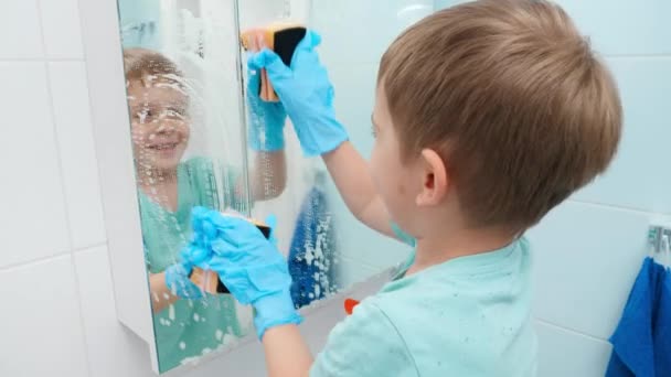 Смешной улыбающийся мальчик стирает зеркало в ванной химическим моющим средством и губками. Дети помогают родителям в повседневной жизни и уборке дома — стоковое видео
