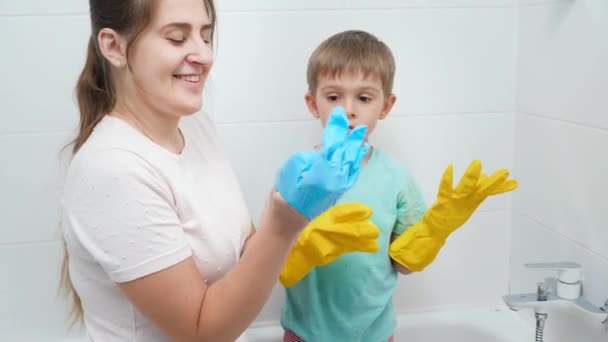 Portret uśmiechniętej i śmiejącej się matki z małym synkiem zakładającym ochronne gumowe rękawiczki przed sprzątaniem domu i myciem łazienki — Wideo stockowe