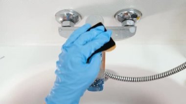 Mavi kauçuk eldivenli bir kadının su musluğunu yıkayıp cilalarkenki görüntüsü. Ev hanımı musluğu sünger ve kimyasal deterjanla temizliyor.