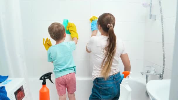 Ung mor med sin lilla son som har roligt och ler när hon tvättar väggar i badrummet. Familjen njuter av hushållsarbete och städning i hemmet — Stockvideo