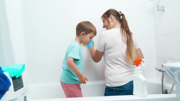 Divertido niño bailando y divirtiéndose mientras madre haciendo tareas domésticas y lavando paredes en el baño — Vídeo de stock