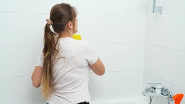 Zadní pohled na mladou ženu v gumových rukavicích mytí stěn v koupelně s houbou a chemickým čisticím prostředkem — Stock video