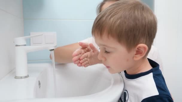 Портрет улыбающегося мальчика с матерью, которая моет и чистит грязный рот водой в раковине ванной комнаты. Детская стирка. — стоковое видео