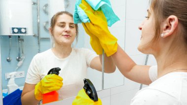 Gülümseyen genç kadın banyo aynasını kimyasal deterjan spreyiyle temizleyip yıkıyor. Ev hanımı ev temizliği ve ev işleri yapıyor.
