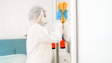 Koruyucu tıbbi giysi, maske ve eldiven giyen ev hanımı evi virüslerden ve bakterilerden arındırıyor. İnsanlar covid-19 ve koronavirüs salgınından korkuyorlar.