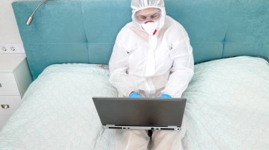 Tıbbi maskeli ve koruyucu giysili bir kadın yatağa uzanıyor ve dizüstü bilgisayarla çalışıyor. Küresel salgın ve covid-19 sırasında uzaktan çalışma