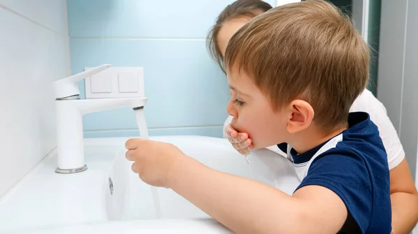 Mosolygó kisfiú arcképe, anya mossa és mossa a mocskos száját vízzel a mosdóban. Gyermekmosás. — Stock Fotó