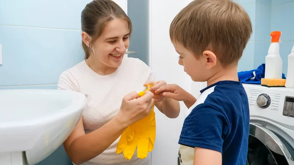 Lächelnde junge Mutter, die ihrem kleinen Sohn hilft, gelbe Gummihandschuhe anzuziehen, bevor sie im Badezimmer Hausarbeit erledigt. Kinder helfen Eltern bei Hausarbeit und Alltag. — Stockfoto