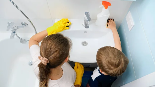 Visão traseira da mãe com filho pequeno limpeza e lavagem de pia de água e torneira no banheiro. Família fazendo tarefas domésticas e tarefas — Fotografia de Stock