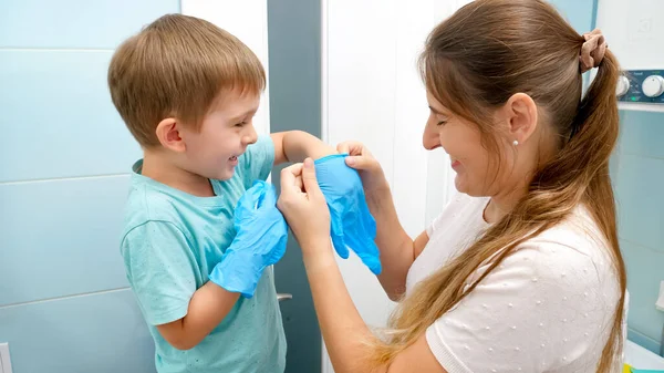 Madre sorridente che aiuta il suo piccolo figlio a indossare e indossare guanti di gomma protettivi prima di fare pulizia della casa.Famiglia che fa i lavori domestici e si diverte — Foto Stock