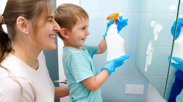 Feliz madre sonriente con hijo pequeño aplicando detergente químico para lavar y limpiar espejos en el baño. Familia haciendo la rutina diaria y tareas domésticas — Foto de Stock