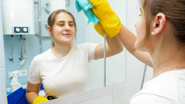 Mujer joven sonriente rociando limpiador químico o detergente en el espejo para limpiar un pulido. Ama de casa en la rutina diaria y las tareas domésticas — Foto de Stock