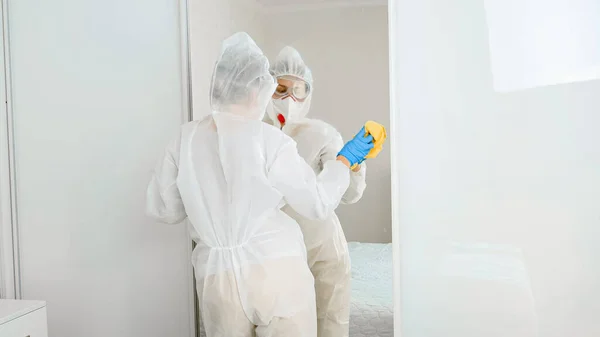 Giovane donna in tuta protettiva medica e maschera respiratore disinfezione mobili e pulizia grande specchio a casa. Persone che combattono e temono di covid-19 e coronavirus — Foto Stock