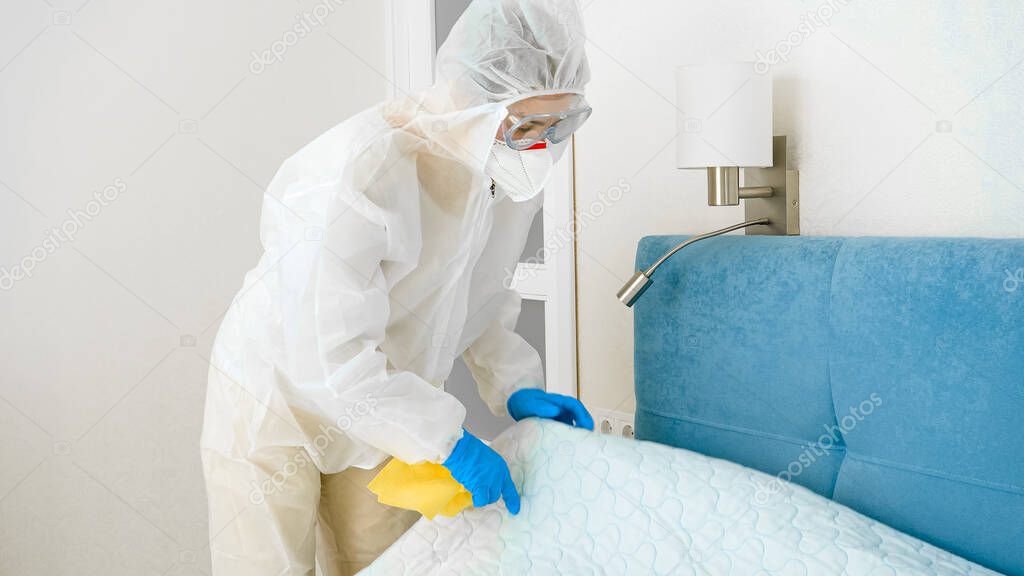 Housewife in protective medical suit and mask doing cleanup and making bed at home