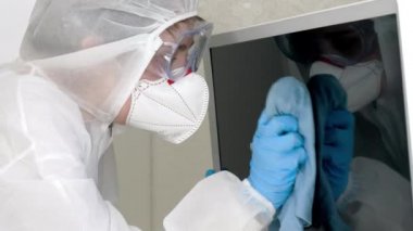 Tıbbi takım elbiseli ve koruyucu maskeli bir kadının ev işi yapan ve evdeki televizyon ve mobilyaları dezenfekte eden yakın plan portresi. Covid-19 fobisi ve koronavirüs paniği