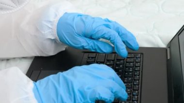 Laptop klavyesinde yazan koruyucu tıbbi eldiven giyen ellerin yakın çekimi. Kilitleme sırasında uzaktan ev oluşturma çalışması