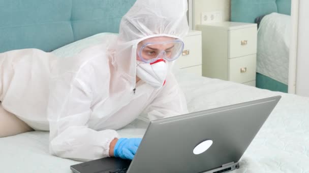 Potret pekerja wanita mengenakan pakaian medis pelindung dan masker wajah berbaring di tempat tidur dan bekerja pada komputer laptop. Orang-orang takut pandemi covid-19 dan coronavirus tinggal di rumah selama kuncian — Stok Video