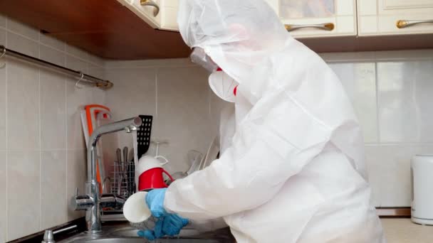 Biyolojik tehlike elbiseli bir kadın mutfakta bulaşık yıkıyor. İnsanlar koronavirüsten ve covid-19 'un evde kalmasından korkuyorlar. — Stok video