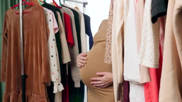 Беременная молодая женщина ходит между длинными стеллажами одежды на вешалках в торговом центре или бутике — стоковое видео