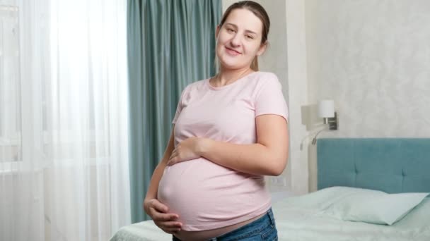 Porträtt av vacker gravid kvinna med stor mage poserar vid fönstret i sovrummet och tittar i kameran — Stockvideo