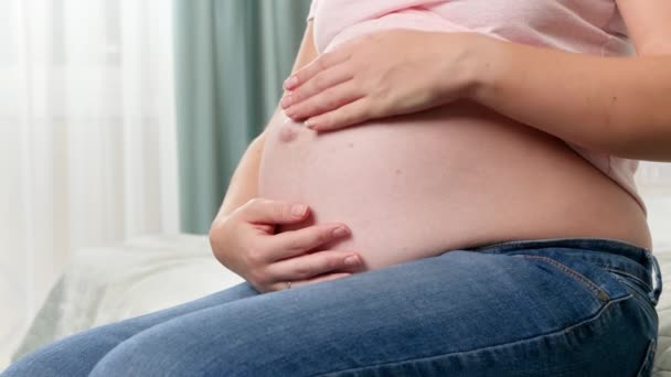 Close-up van mooie zwangere vrouw met grote buik zitten op bed en aanraken van haar buik met handen — Stockvideo