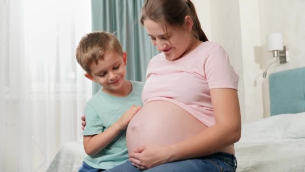 Lindo niño tocando gran vientre de su madre embarazada sentada en la cama en el dormitorio. Emociones positivas en la familia esperando al bebé — Vídeos de Stock