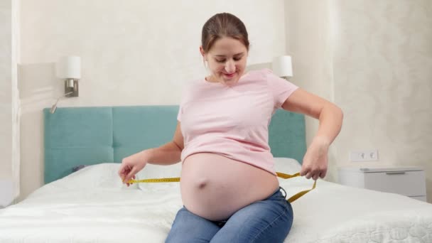 Gelukkig glimlachen zwangere vrouw zitten op bed en het meten van haar grote buik met meetlint — Stockvideo